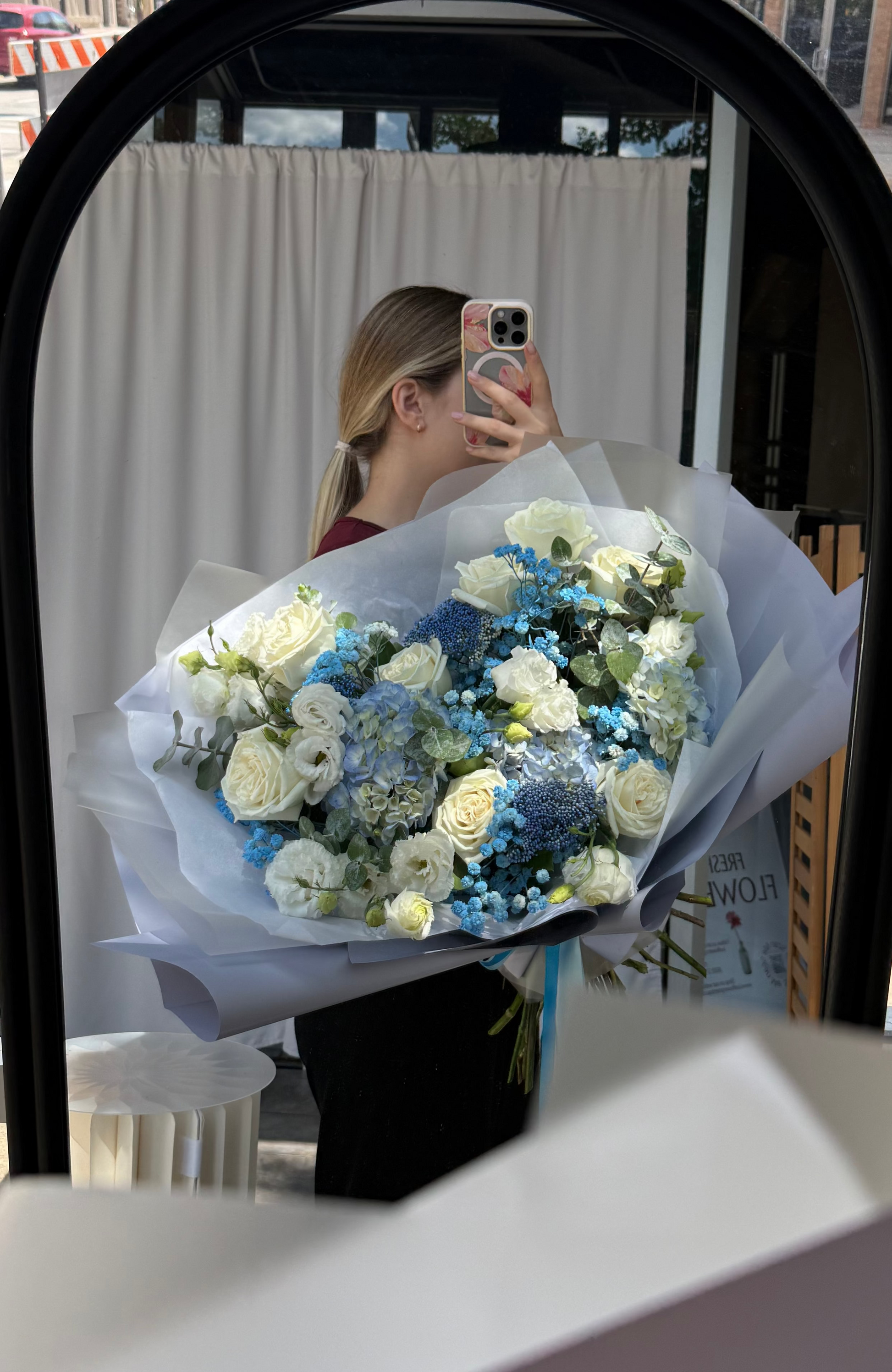 A person taking a mirror selfie in a storefront, holding a bouquet of white and blue flowers wrapped in light gray paper. The scene is reflected in a black-framed oval mirror, with a city street and pedestrians visible outside.