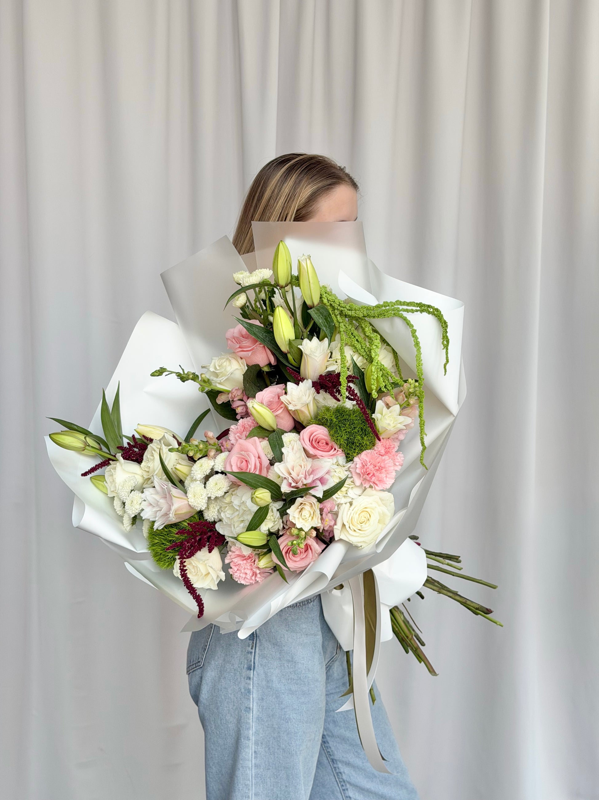 A person holding a large, beautifully wrapped bouquet of pink, white, and burgundy flowers with green foliage, standing in front of a neutral white curtain backdrop.