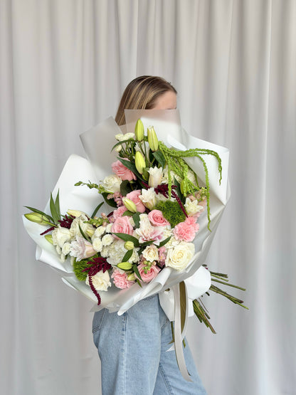 A person holding a large, beautifully wrapped bouquet of pink, white, and burgundy flowers with green foliage, standing in front of a neutral white curtain backdrop.