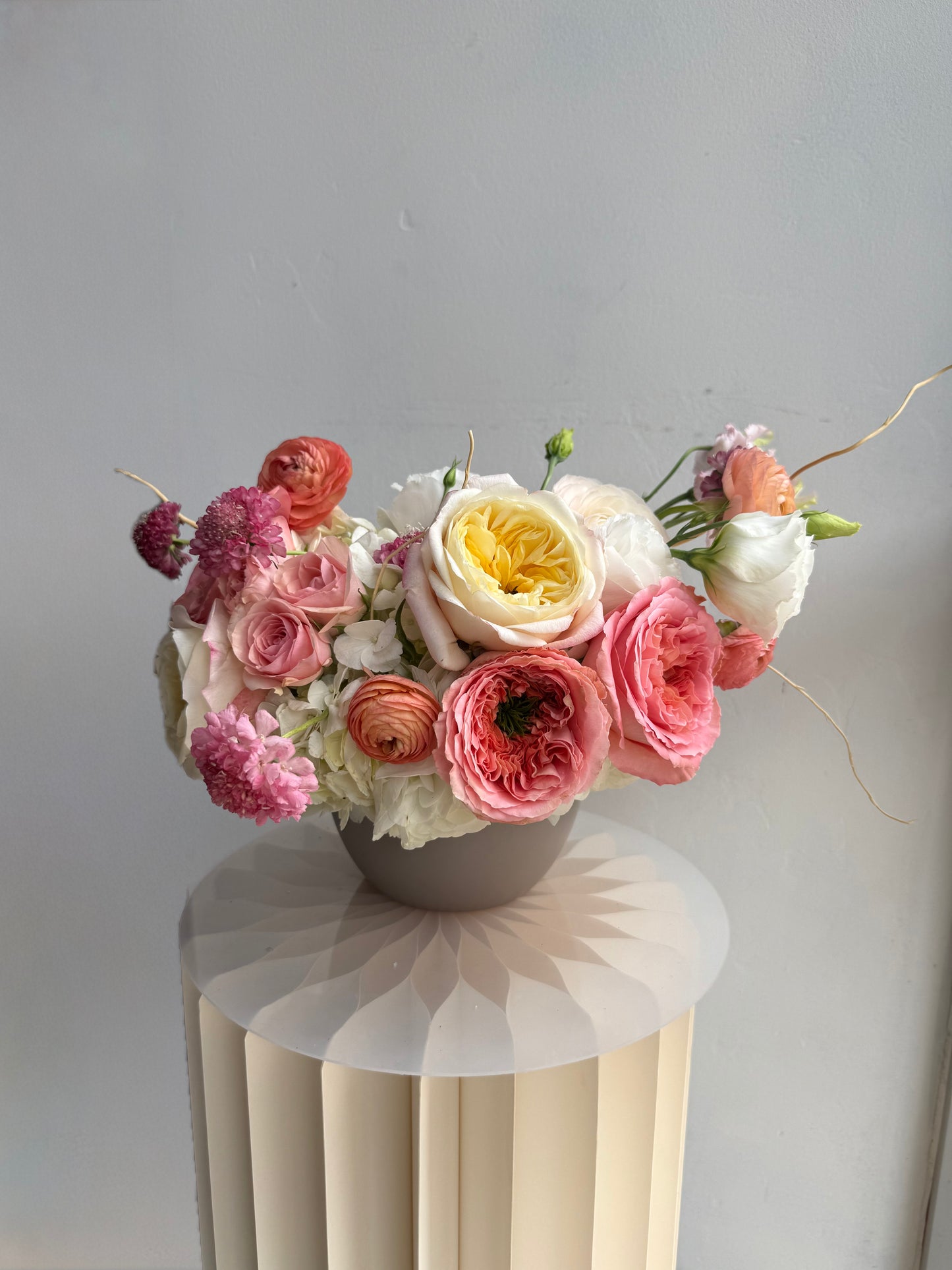A vibrant floral arrangement featuring pink, peach, yellow, and white flowers in a gray pot, displayed on a cream-colored fluted pedestal against a neutral gray wall.