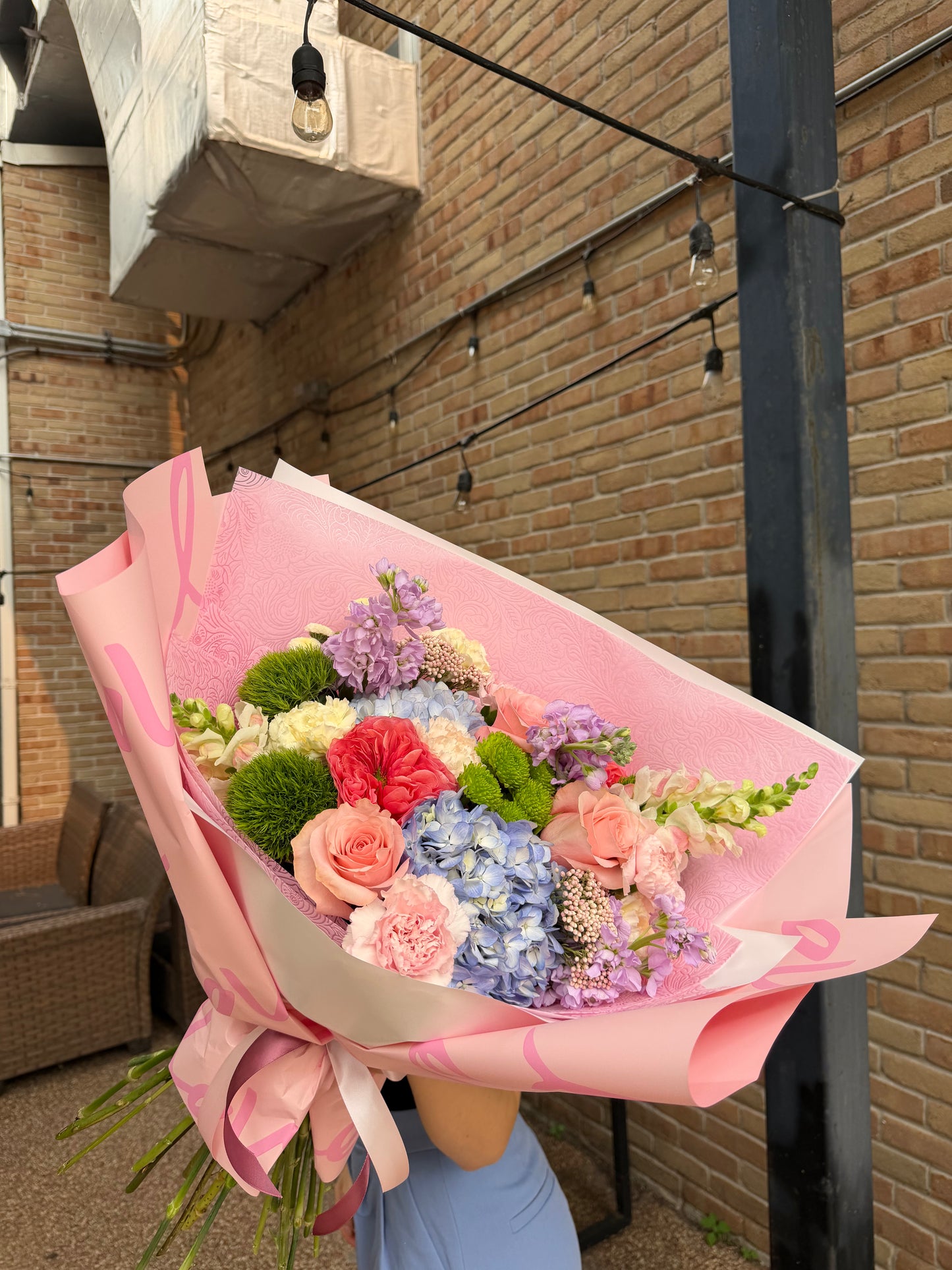 A large, vibrant flower bouquet wrapped in pink paper and held by a person wearing a light blue dress, set against a brick patio background.