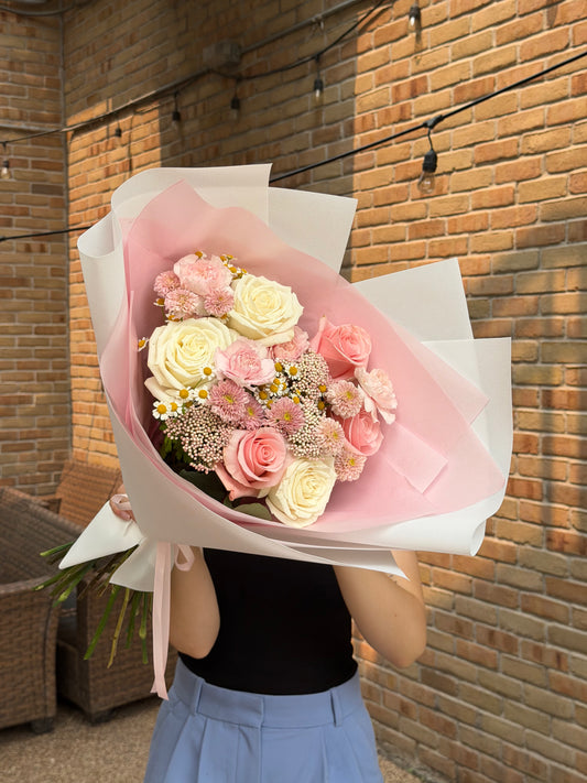 A person holding a large bouquet of pink and cream roses wrapped in white and pink paper, standing in front of a brick wall.